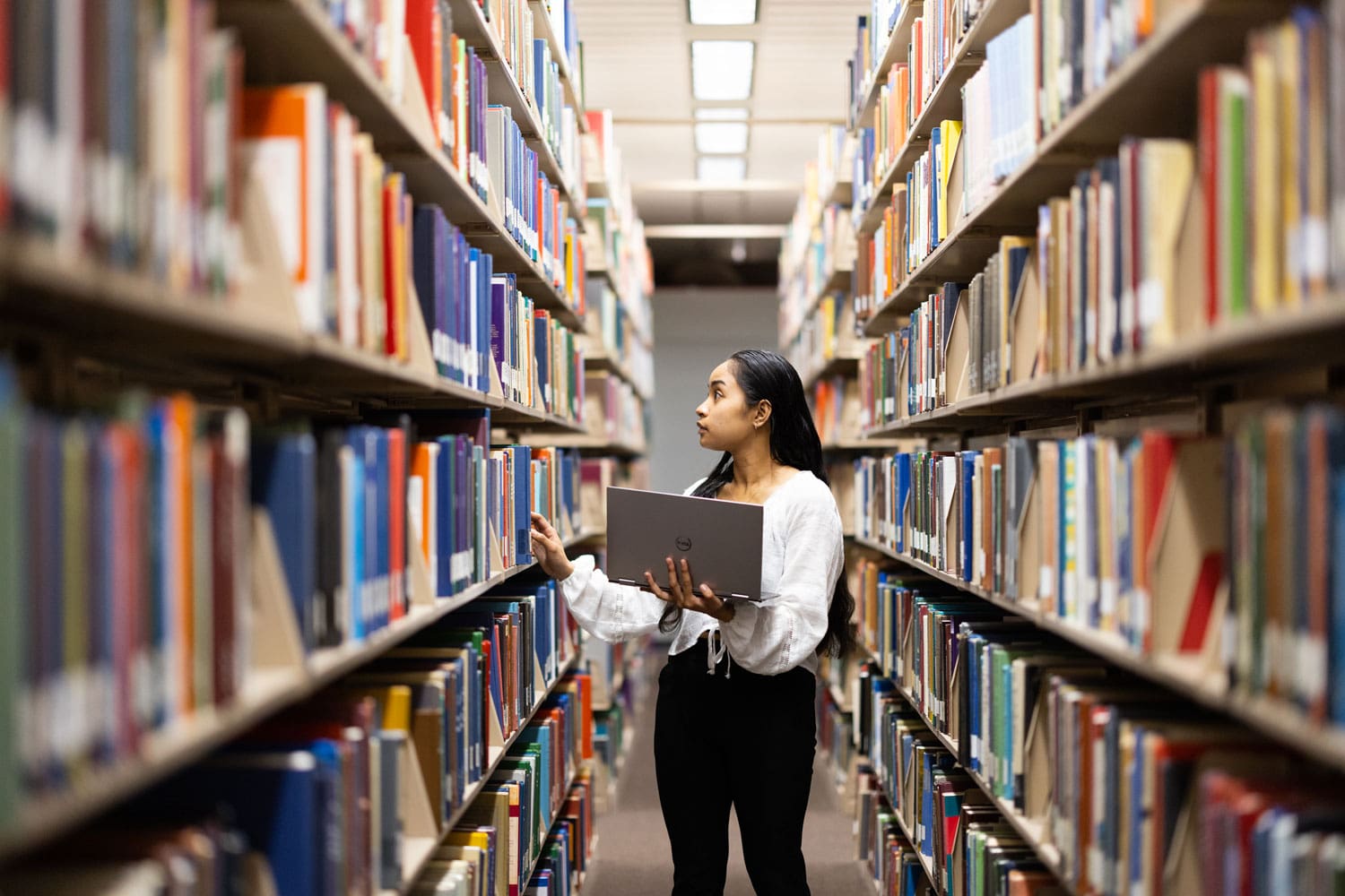 A studenmt holds a laptop while looking through books at the library.